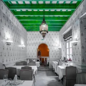 Salón comedor de color blanco con papel estilo neofrances en paredes, techos altos con maderos pintados en verde y una gran lampara estilo arabe en el centro. Mesas a los lados con manteles blancos elegantes y al fondo una pasillo con forma arabe y pared naranja, del restaurante italiano San Marco Santa Cruz en Sevilla
